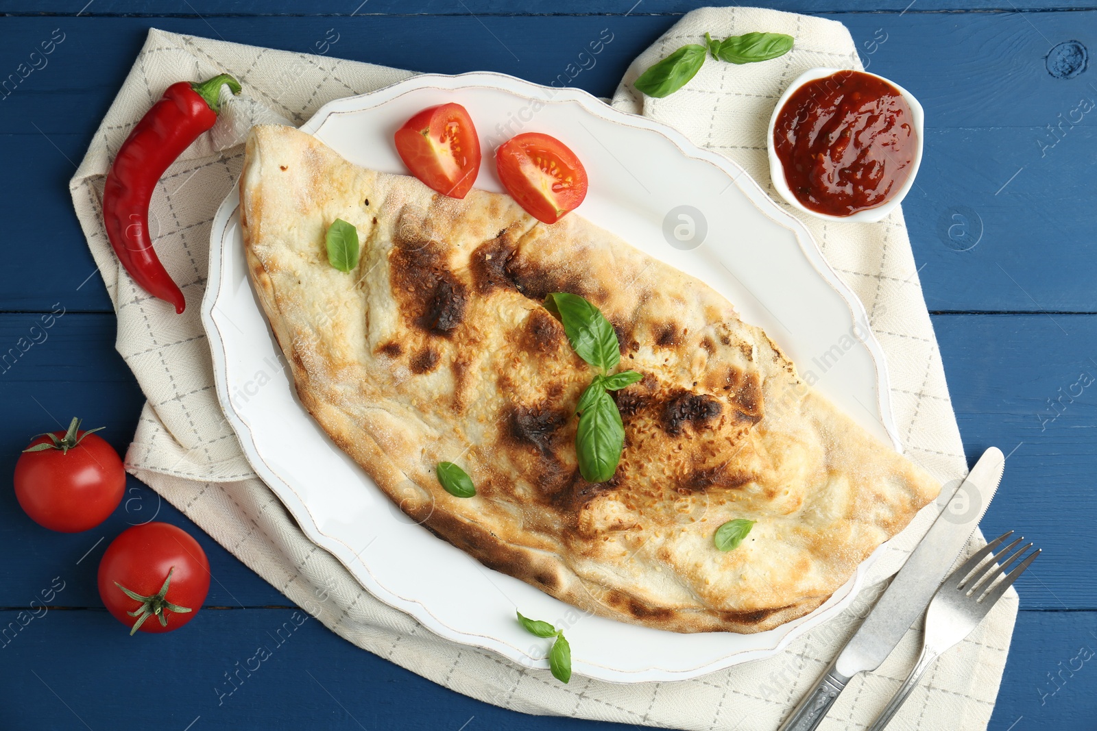 Photo of Tasty calzone with basil, tomatoes, chili pepper and sauce on blue wooden table, flat lay