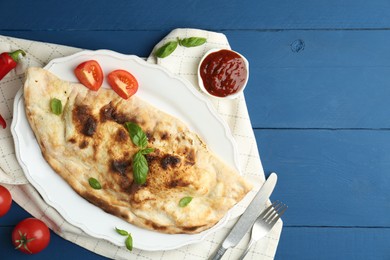 Photo of Tasty calzone with basil, tomatoes and sauce on blue wooden table, flat lay