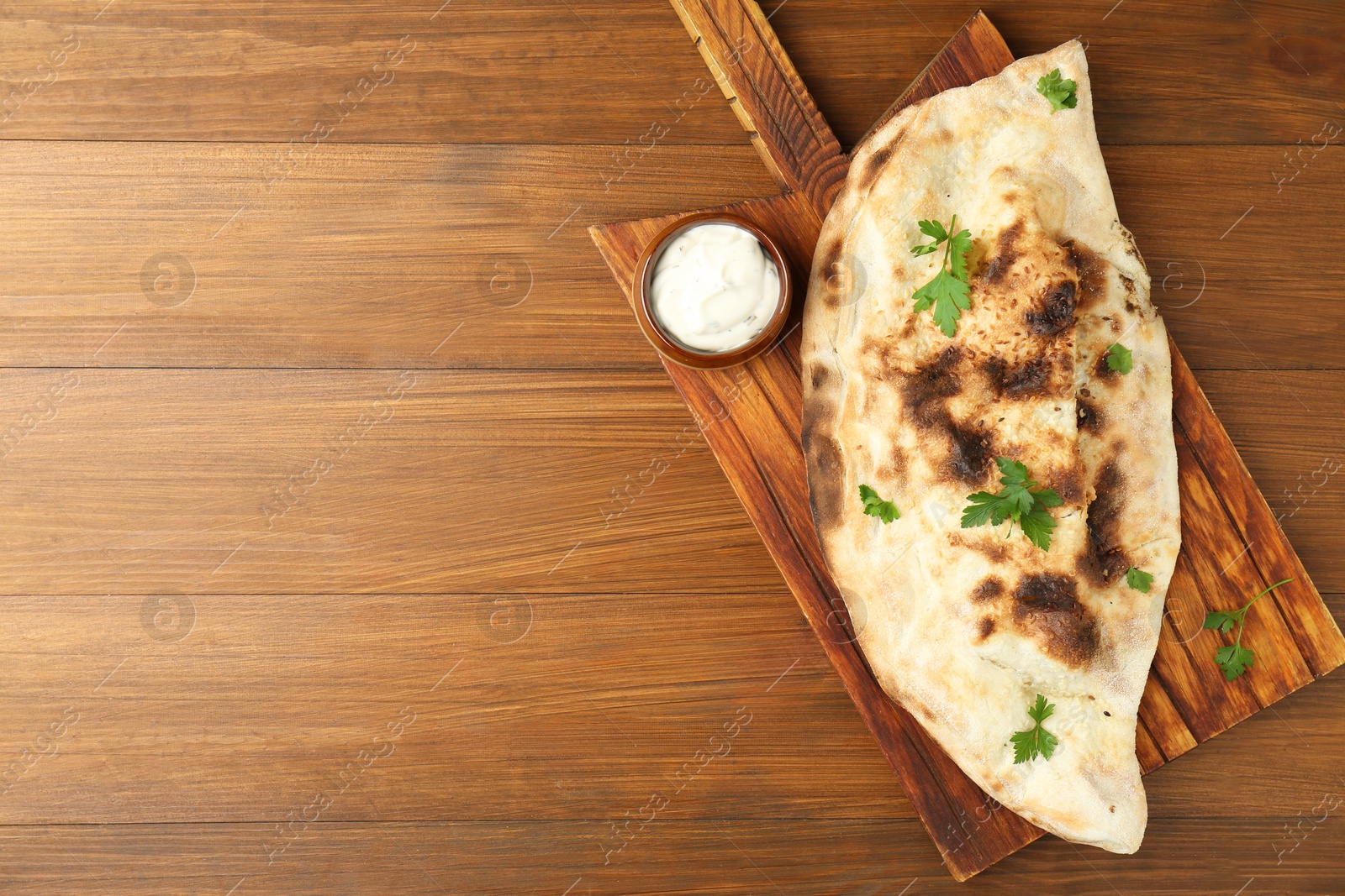 Photo of Board with tasty calzone, parsley and sauce on wooden table, top view. Space for text