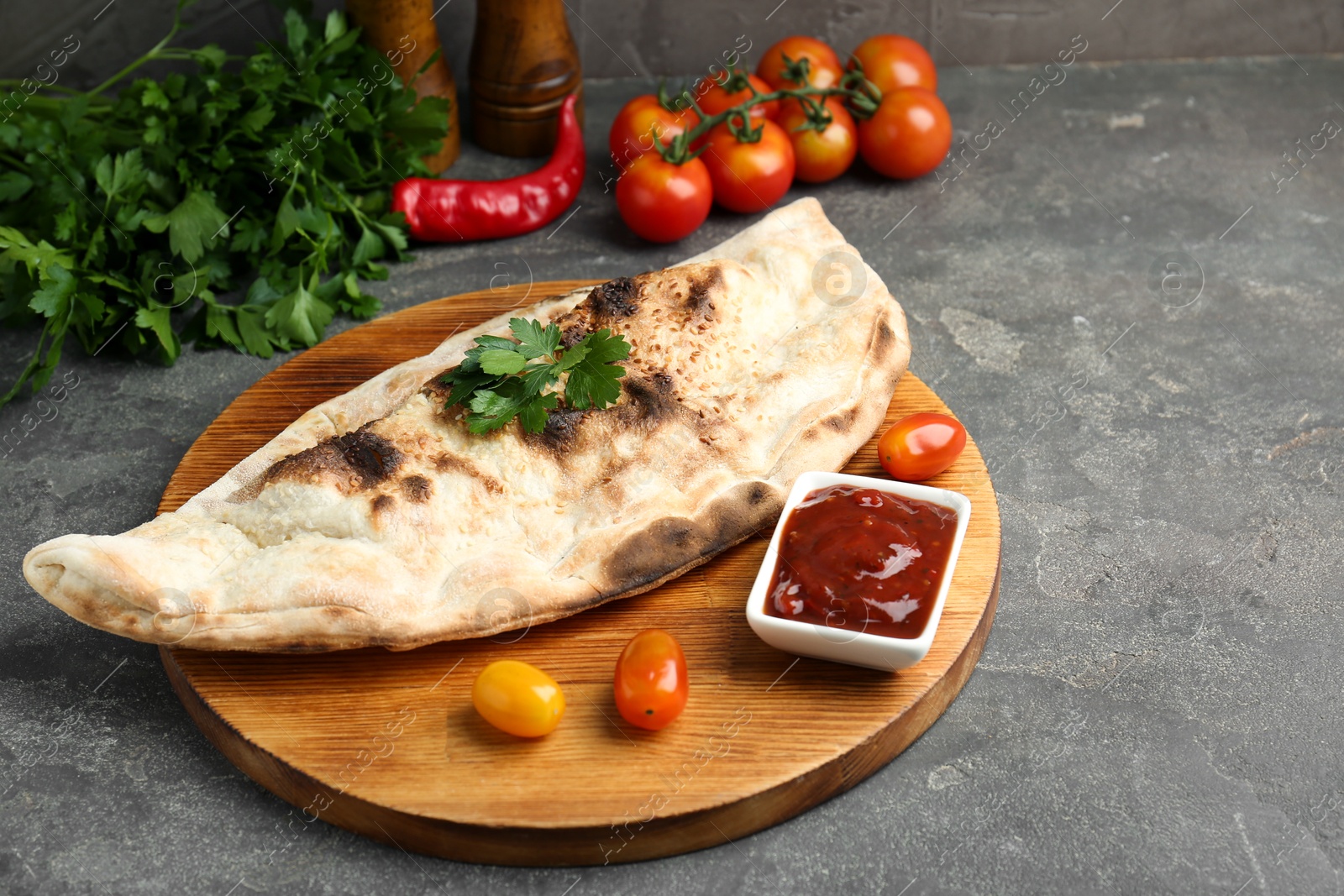 Photo of Board with tasty calzone, tomatoes, sauce and parsley on grey textured table, closeup