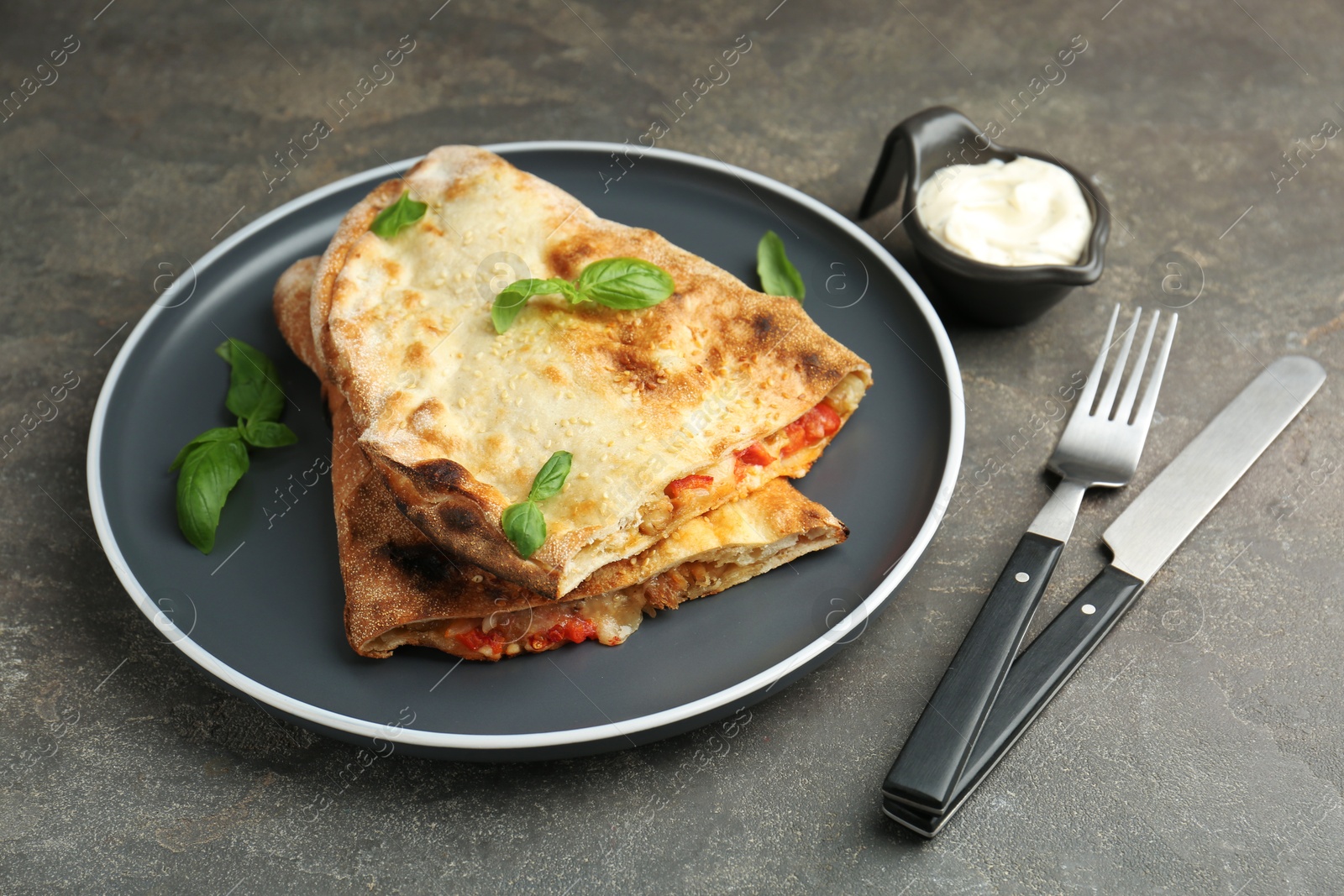 Photo of Halves of tasty calzone with meat, cheese, basil and tomato served with sauce on grey textured table, closeup