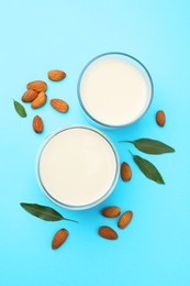 Fresh nut milk in glasses, almonds and green leaves on light blue background, flat lay