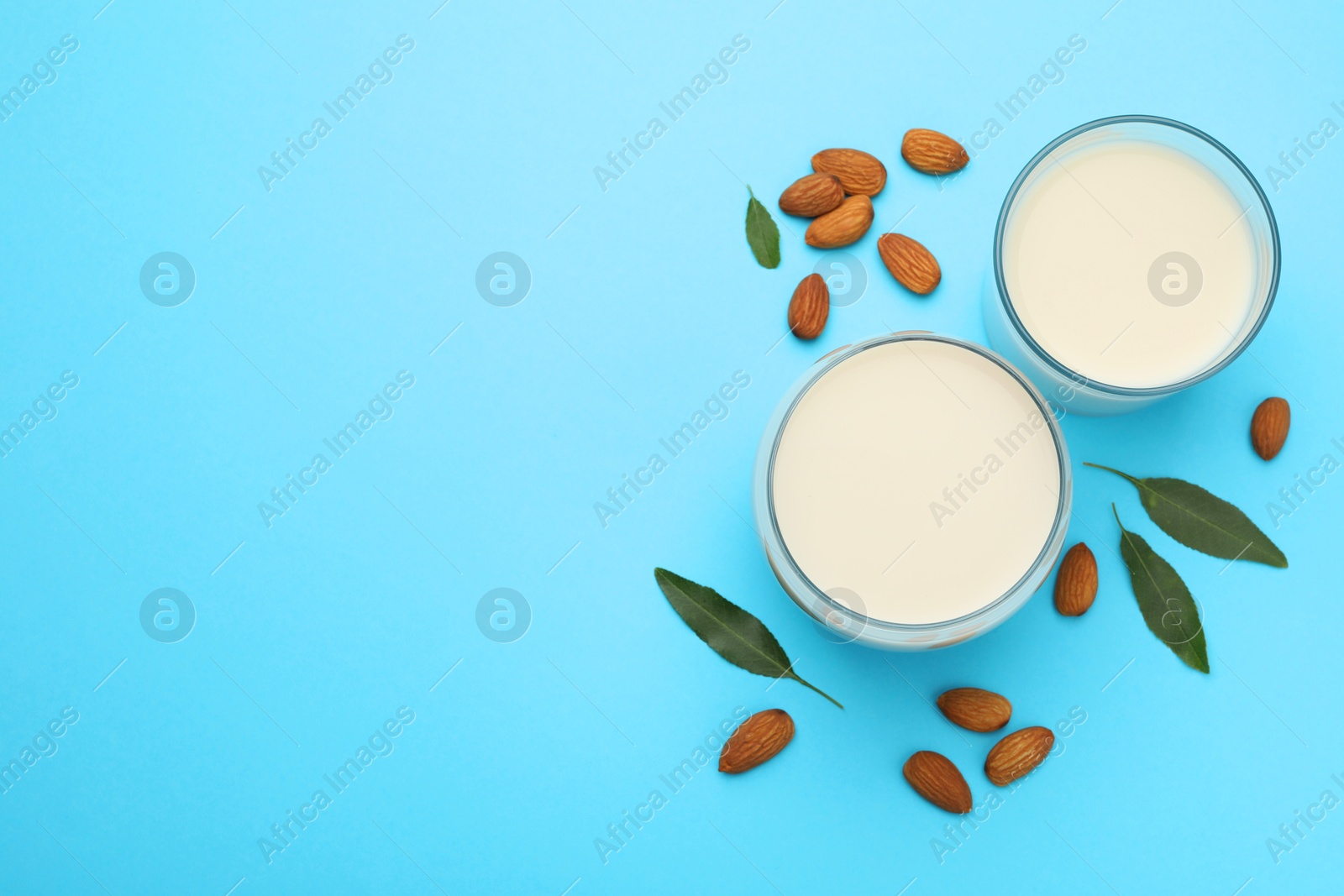 Photo of Fresh nut milk in glasses, almonds and green leaves on light blue background, flat lay. Space for text