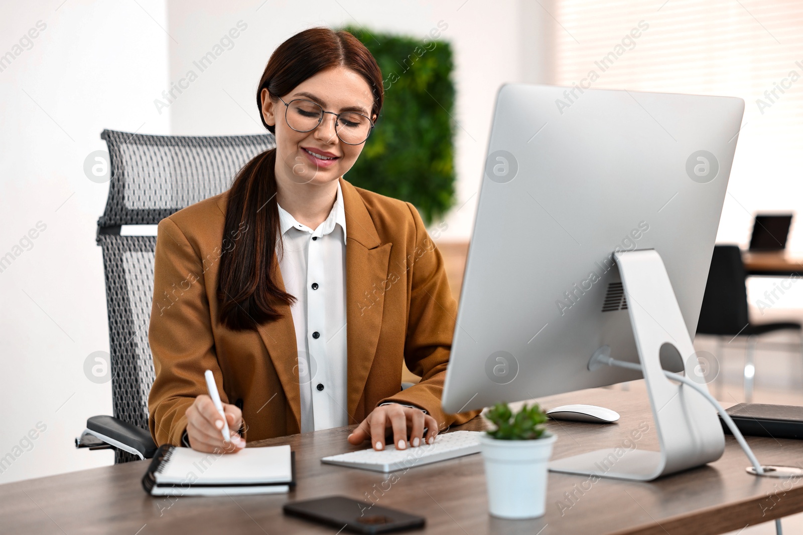 Photo of Woman taking notes while working on computer at table in office