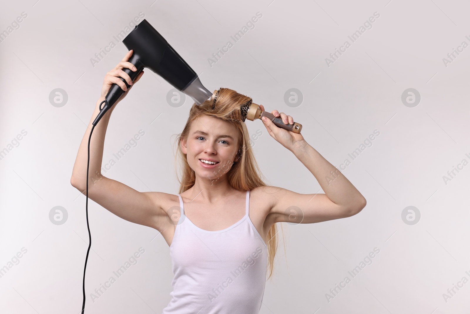 Photo of Beautiful young woman styling her hair with hairdryer and brush on light grey background