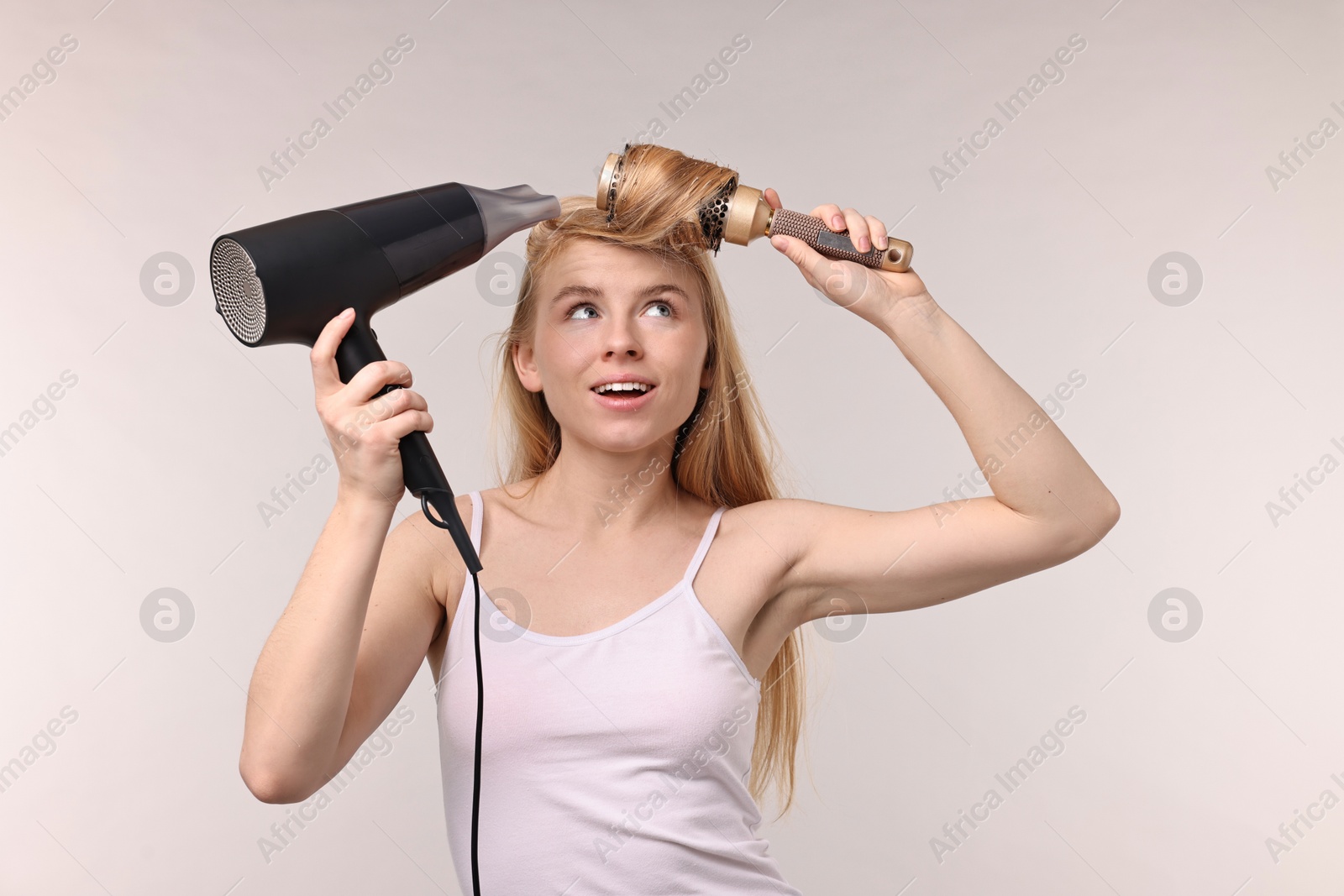 Photo of Beautiful young woman styling her hair with hairdryer and brush on light grey background