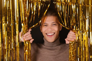 Photo of Happy young man looking out through golden foil curtain against dark background