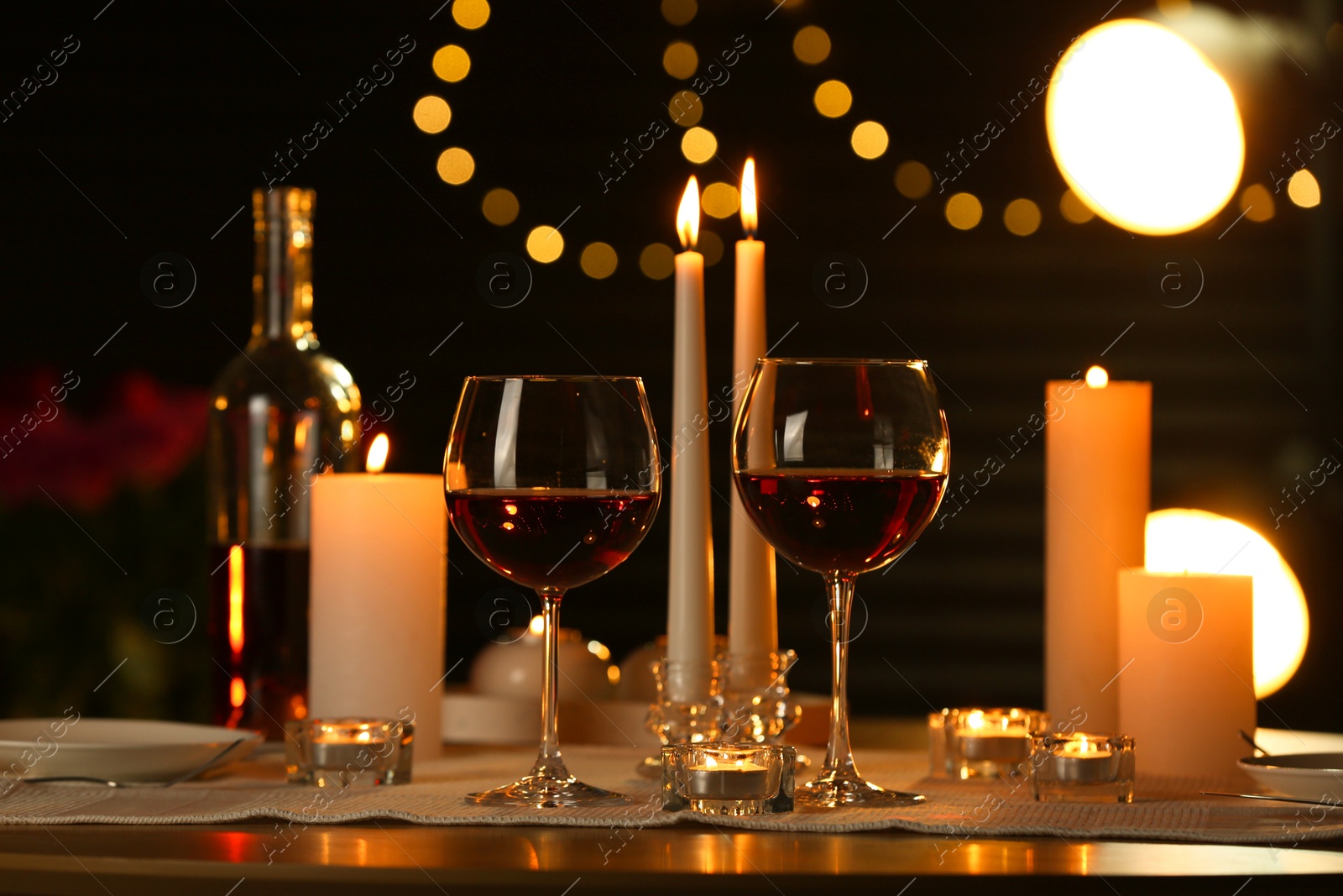 Photo of Romantic table setting with red wine and burning candles against blurred lights