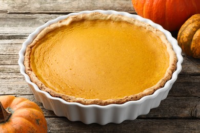 Photo of Tasty homemade pumpkin pie in baking dish and fresh pumpkins on wooden table, closeup