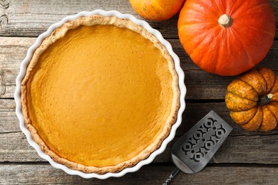 Photo of Tasty homemade pumpkin pie in baking dish, fresh pumpkins and cake server on wooden table, flat lay