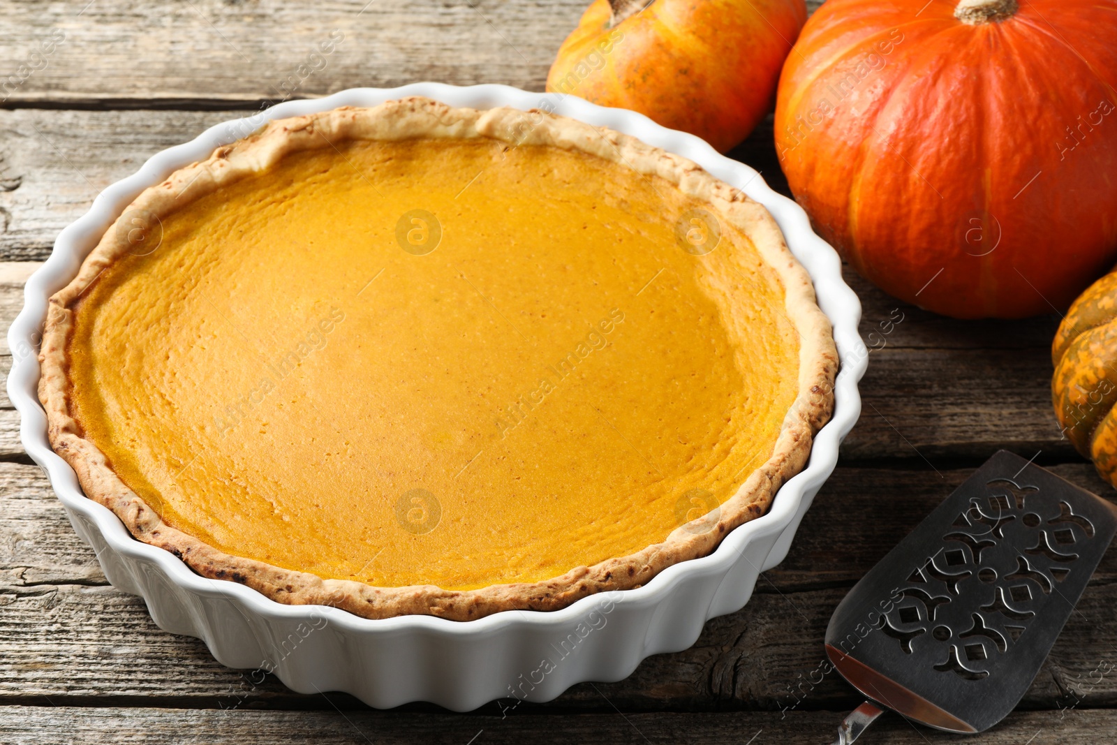 Photo of Tasty homemade pumpkin pie in baking dish, fresh pumpkins and cake server on wooden table, closeup