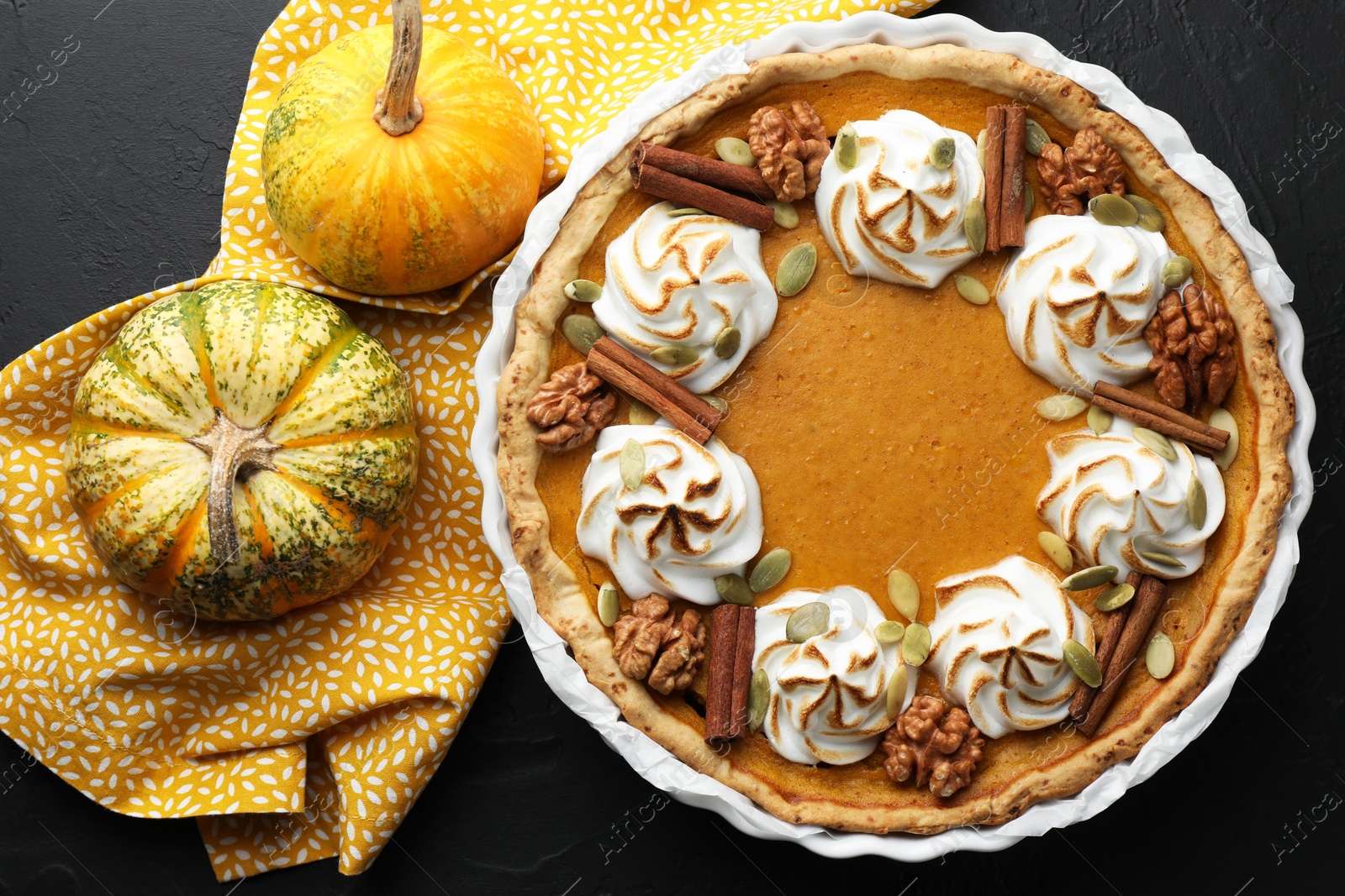 Photo of Delicious homemade pumpkin pie in baking dish and fresh pumpkins on black textured table, flat lay