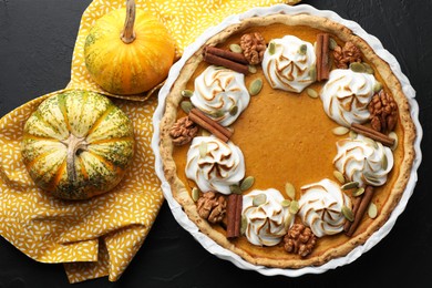 Photo of Delicious homemade pumpkin pie in baking dish and fresh pumpkins on black textured table, flat lay