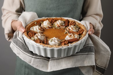 Photo of Woman holding homemade pumpkin pie with whipped cream, seeds and cinnamon on grey background, closeup