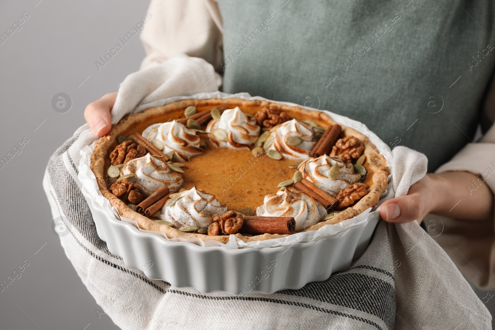 Photo of Woman holding homemade pumpkin pie with whipped cream, seeds and cinnamon on grey background, closeup