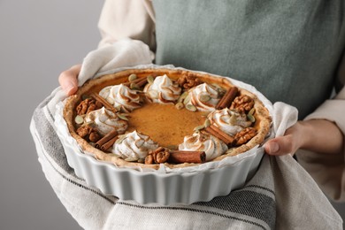 Photo of Woman holding homemade pumpkin pie with whipped cream, seeds and cinnamon on grey background, closeup