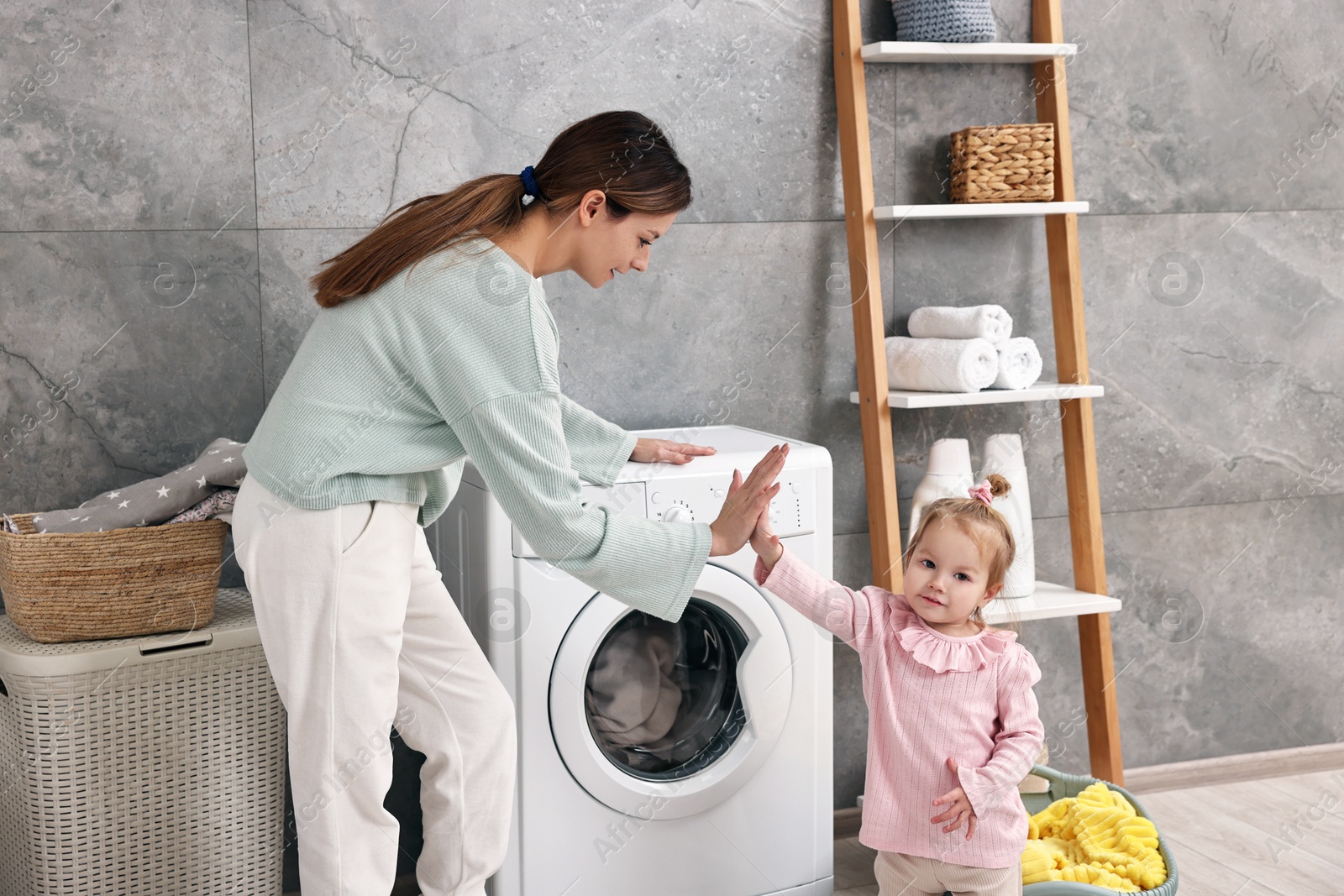 Photo of Smiling housewife giving high five to her little daughter near washing machine at home