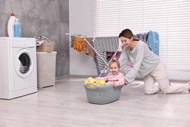 Photo of Smiling housewife and her little daughter having fun with basket of laundry at home