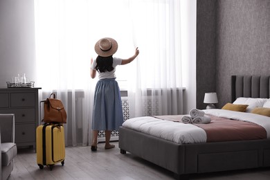 Photo of Traveller with suitcase opening curtains in hotel room, back view