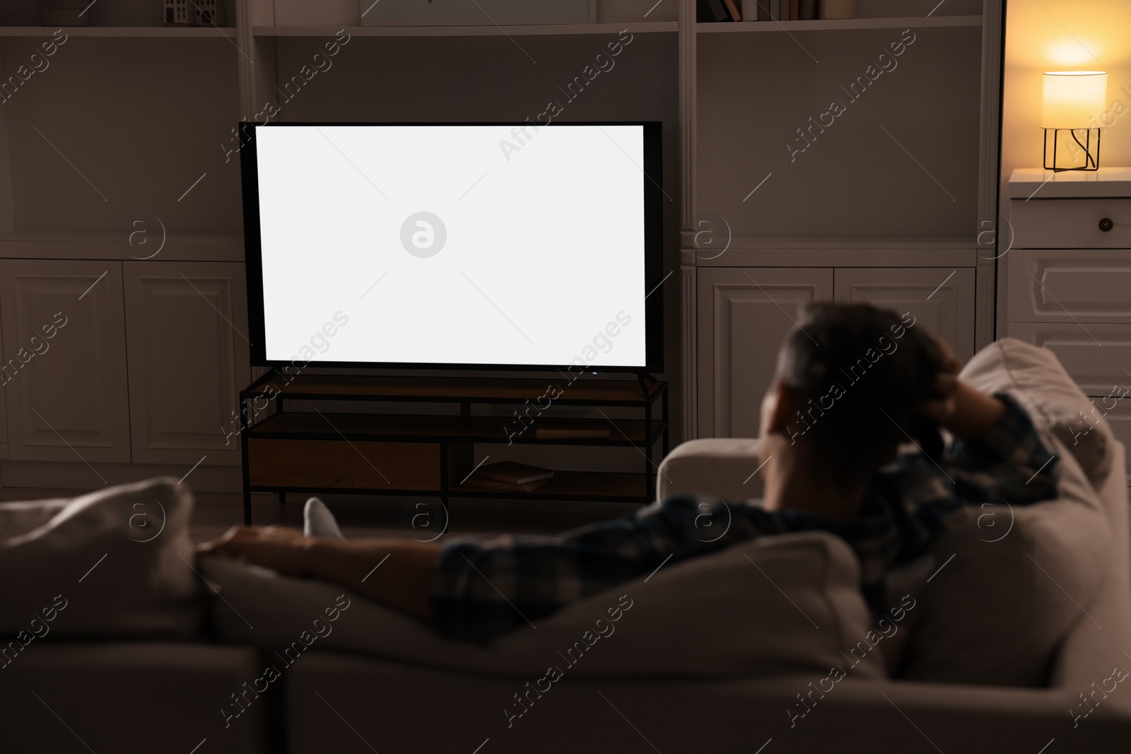 Photo of Man watching TV on sofa at home in evening, back view