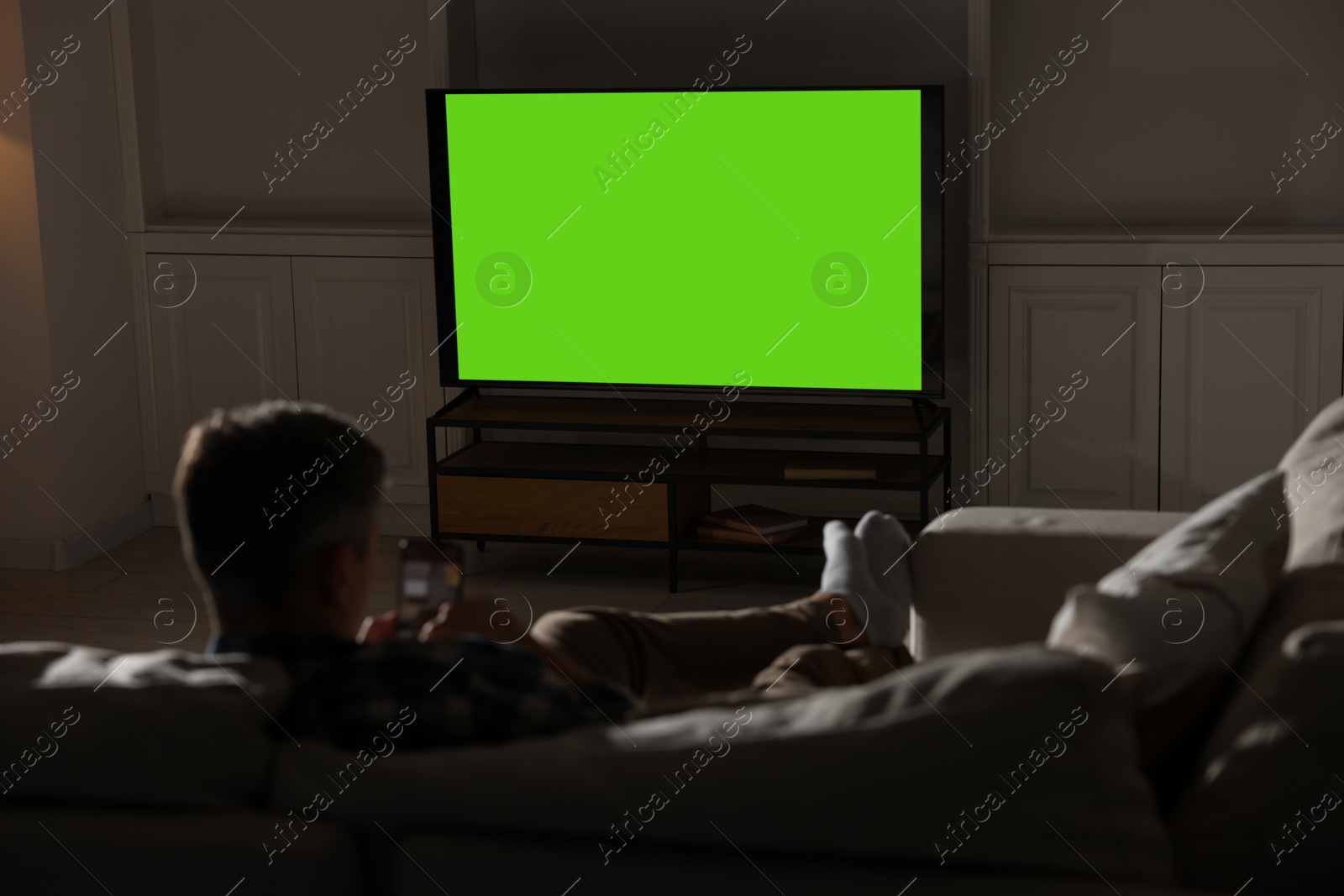 Photo of Man watching TV on sofa at home in evening, back view