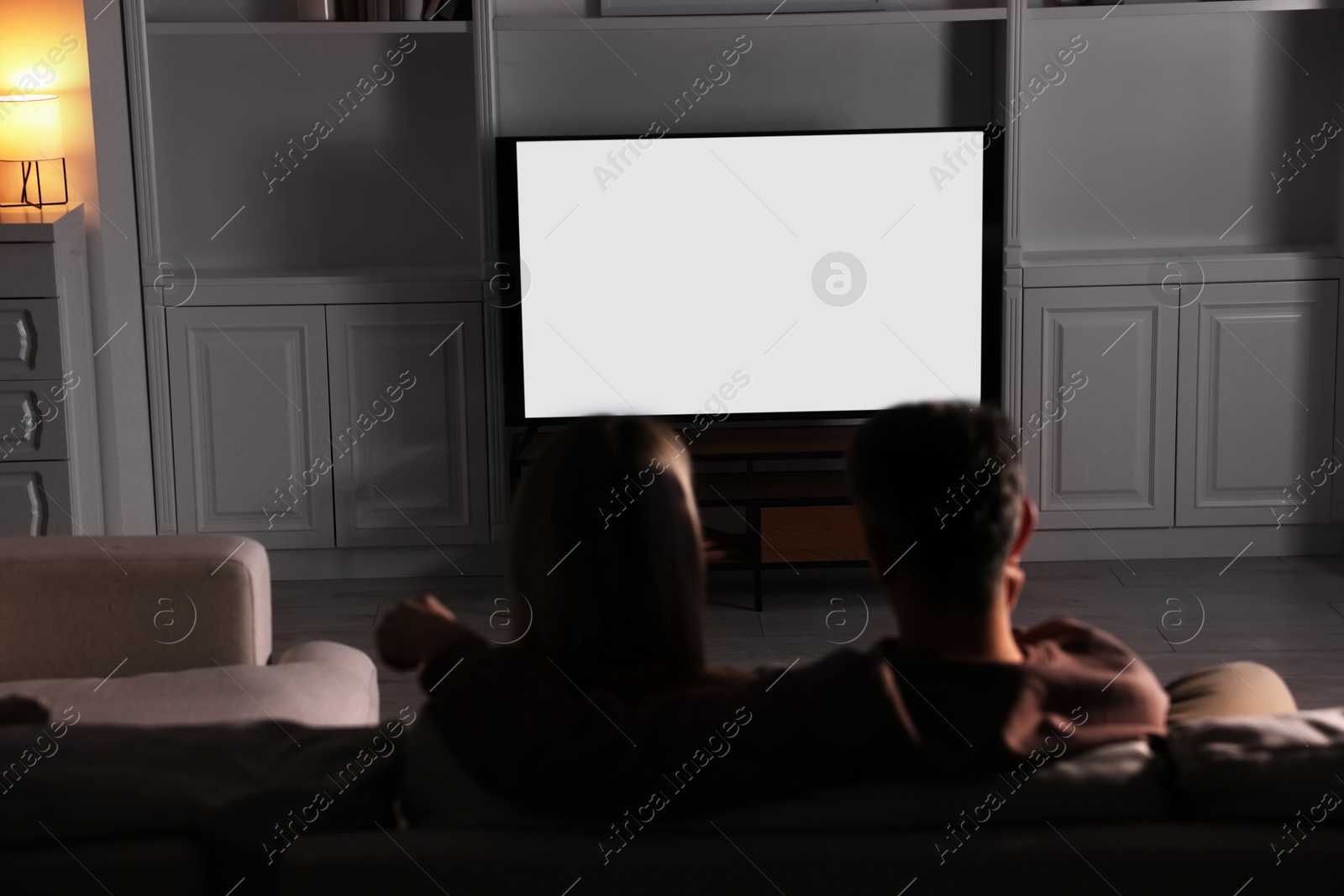 Photo of Couple watching TV on sofa at home in evening, back view