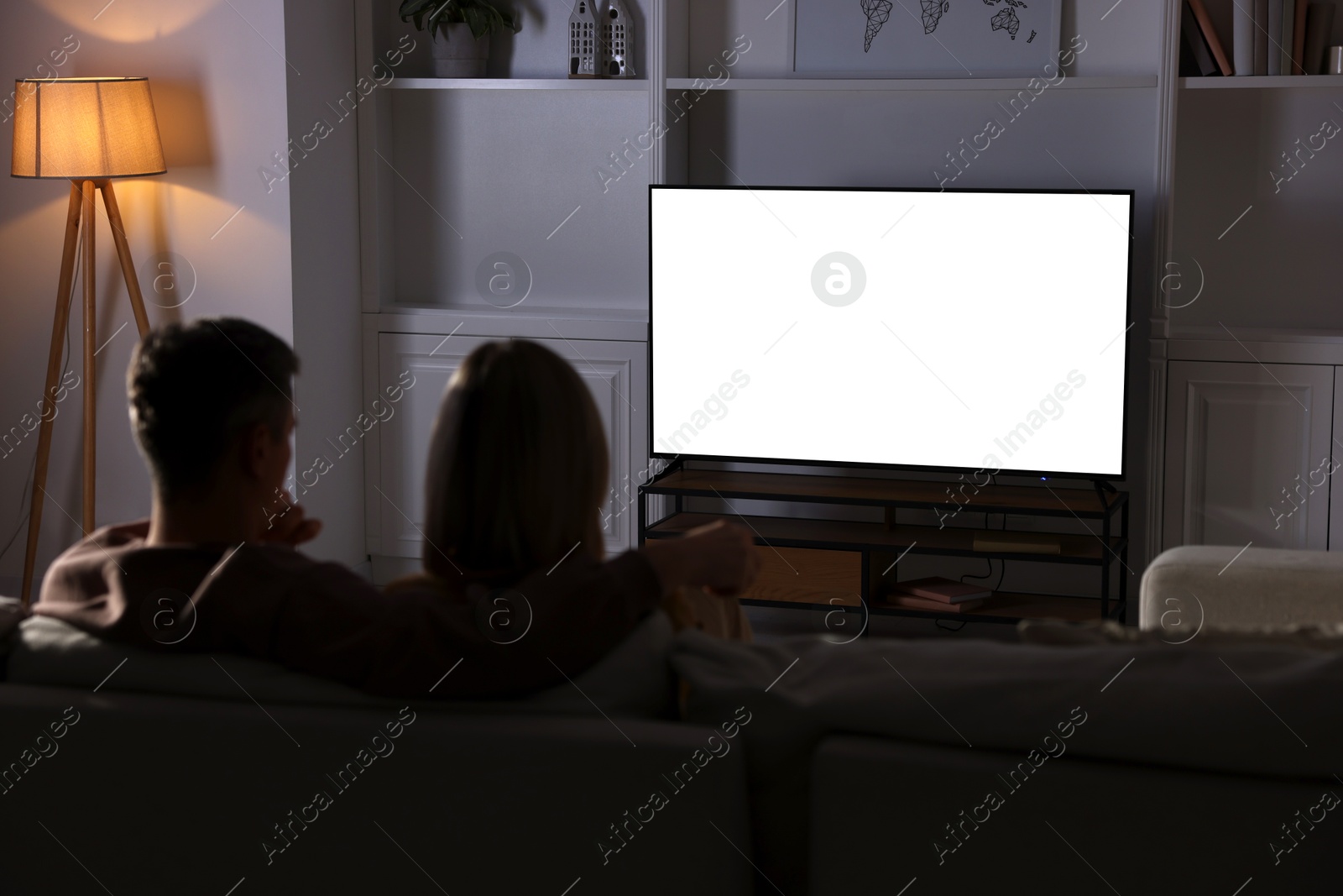 Photo of Couple watching TV on sofa at home in evening, back view