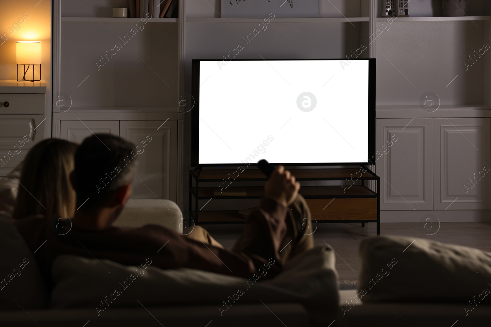 Photo of Couple watching TV on sofa at home in evening, back view