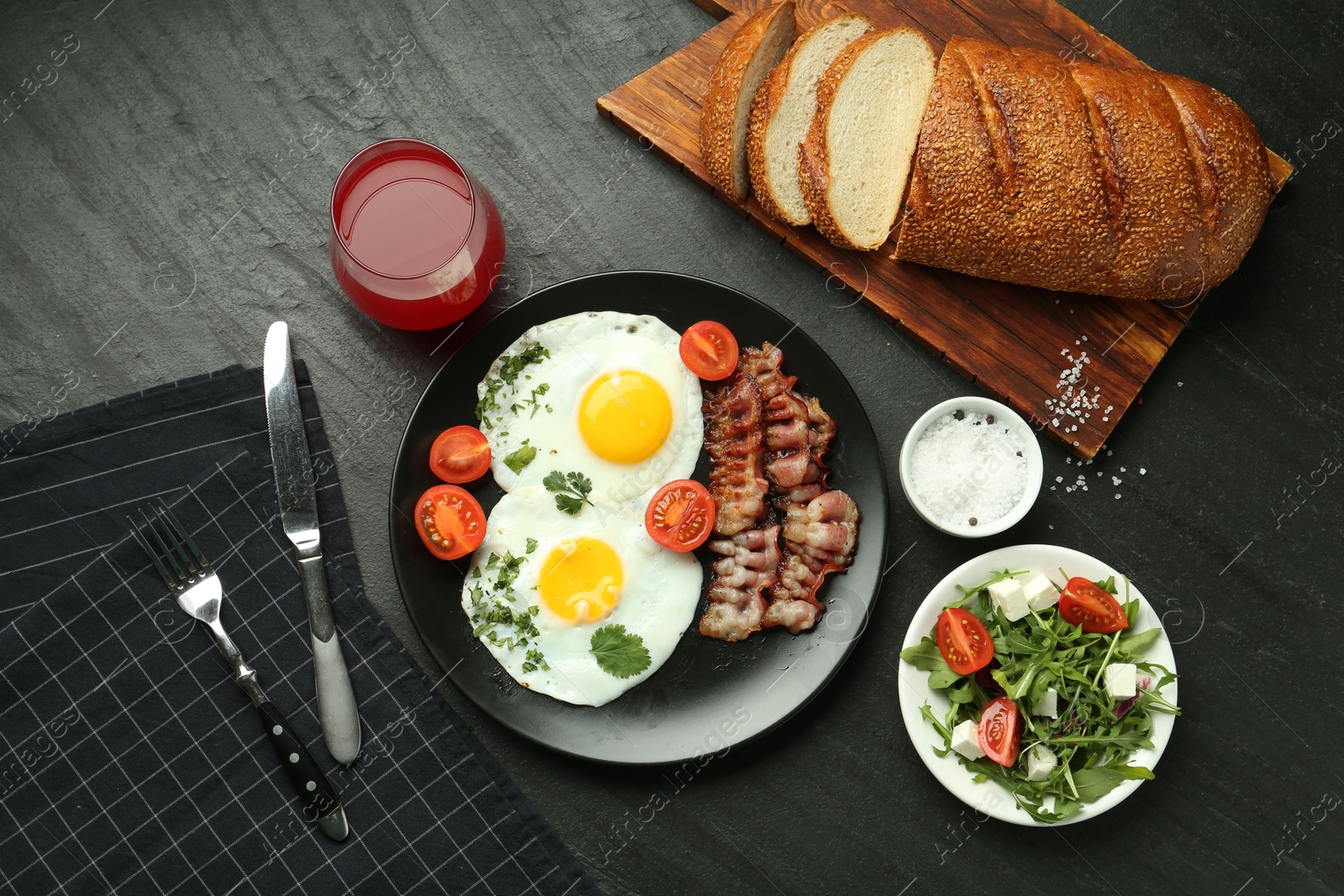 Photo of Tasty brunch. Delicious fried eggs, bacon, salad and glass of juice served on black table, flat lay