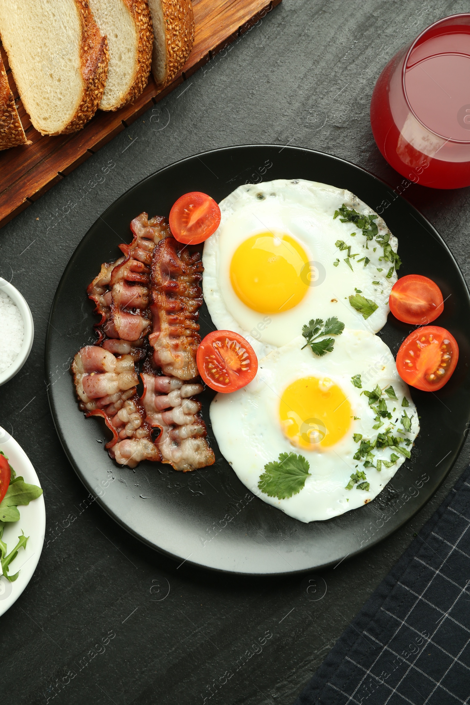 Photo of Tasty brunch. Delicious fried eggs, bacon and glass of juice served on black table, flat lay