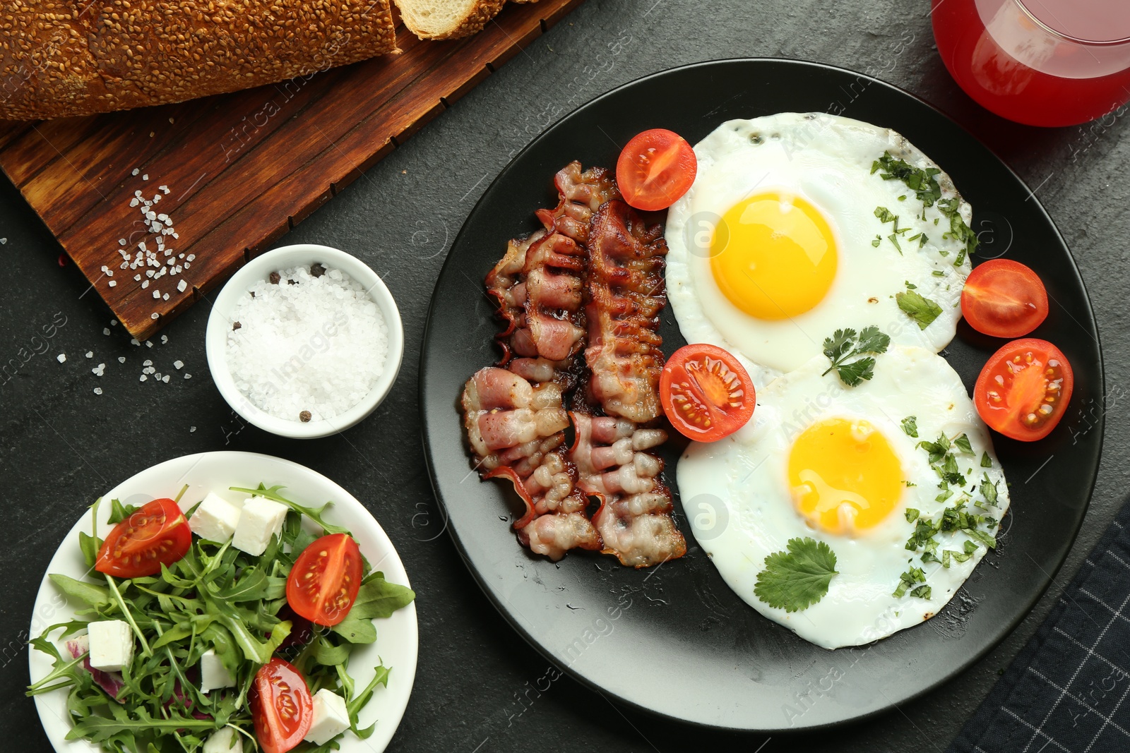 Photo of Tasty brunch. Delicious fried eggs, bacon, salad and glass of juice served on black table, flat lay