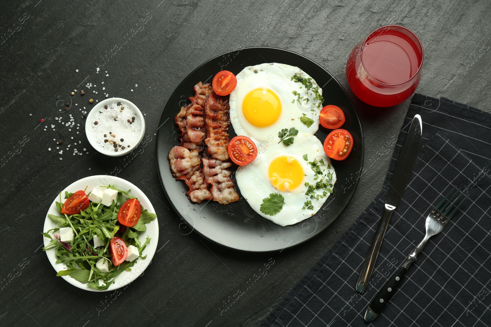 Photo of Tasty brunch. Delicious fried eggs, bacon, salad and glass of juice served on black table, flat lay