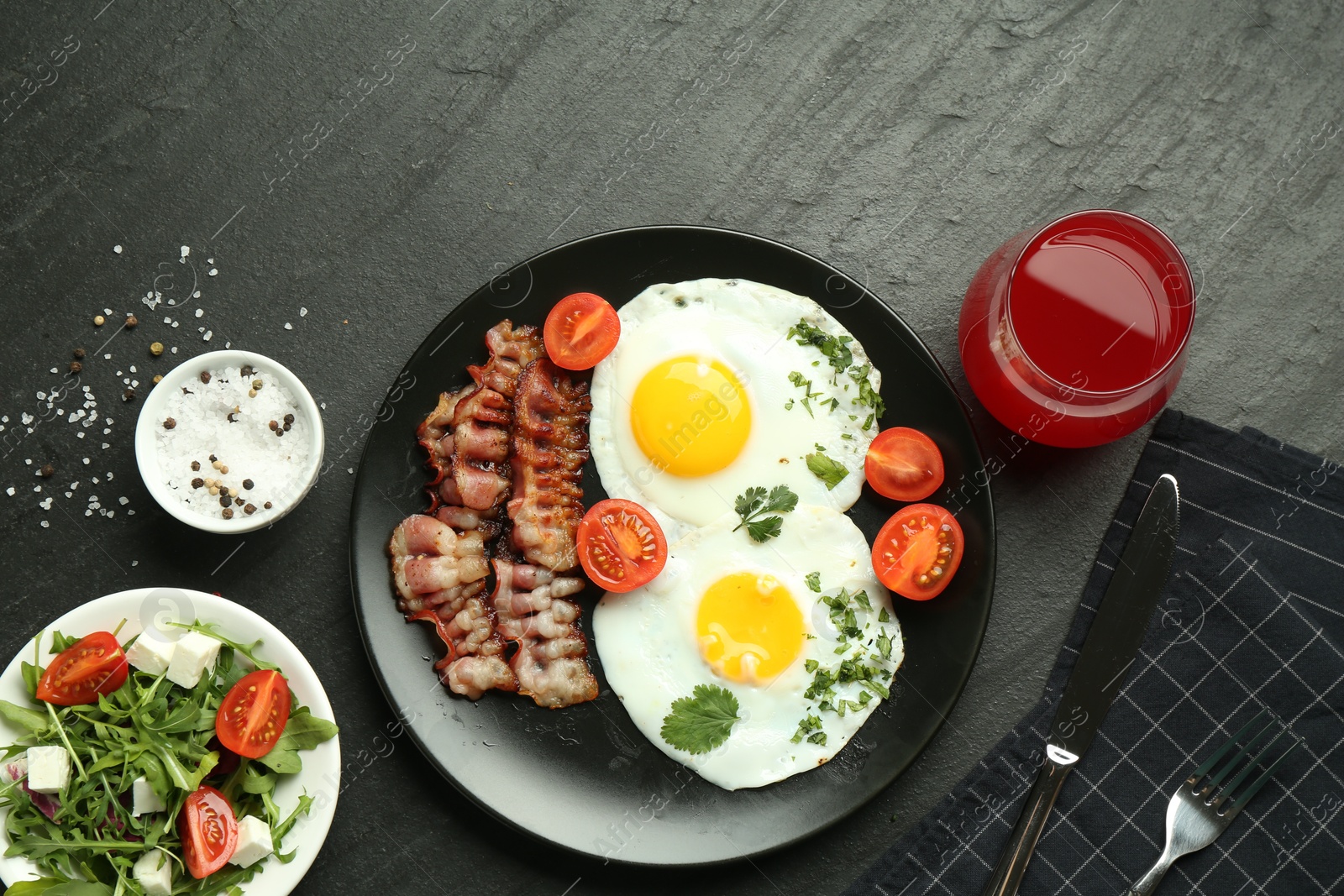 Photo of Tasty brunch. Delicious fried eggs, bacon, salad and glass of juice served on black table, flat lay