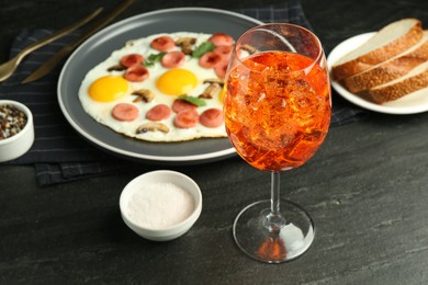 Photo of Tasty brunch. Aperol cocktail, delicious fried eggs with sausage served on black table, closeup