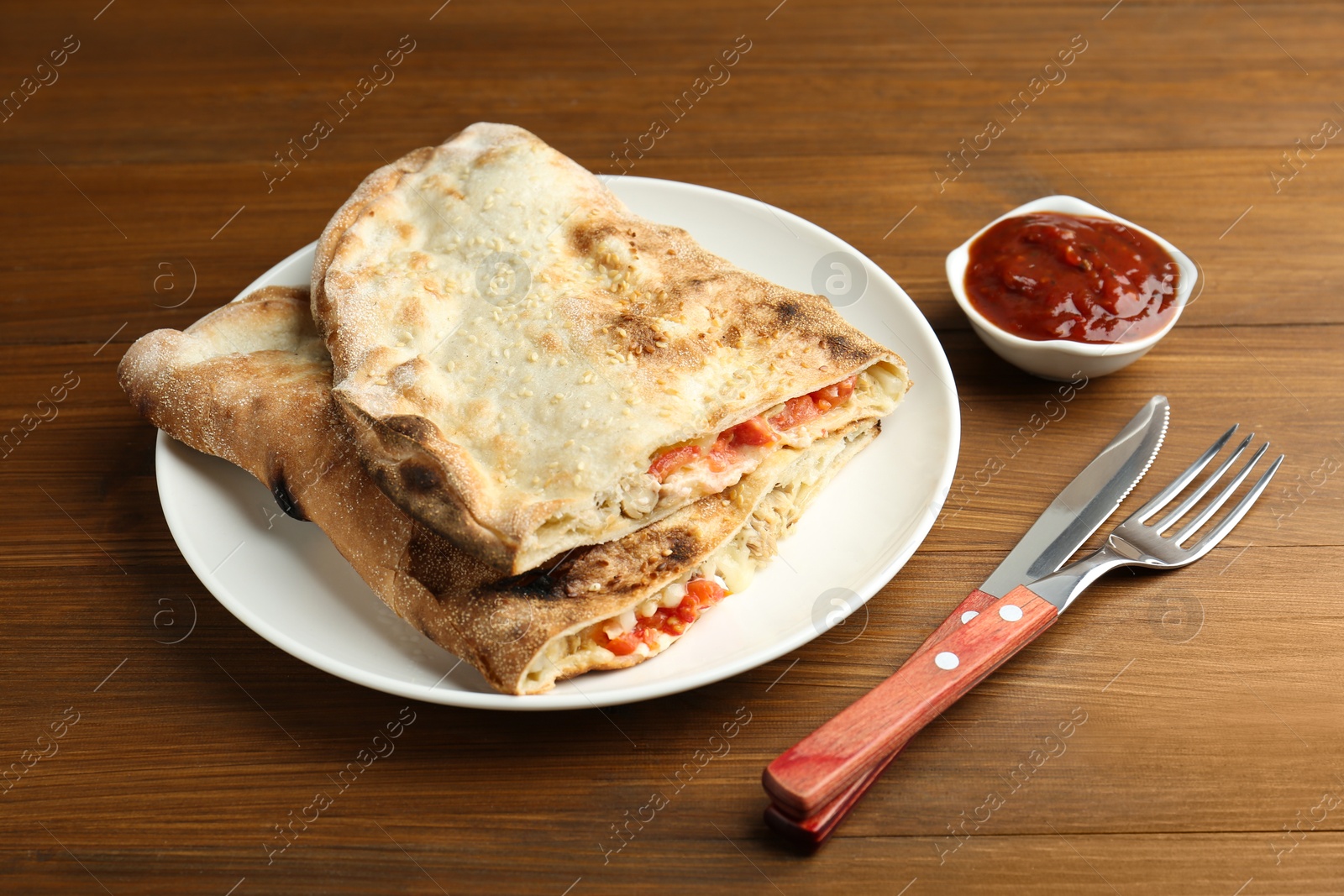 Photo of Halves of tasty calzone with meat, cheese and tomato served with sauce on wooden table, closeup