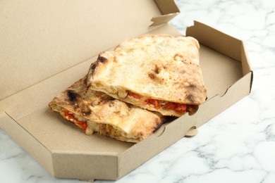 Photo of Halves of tasty calzone with meat, cheese and tomato in cardboard box on white marble table, closeup