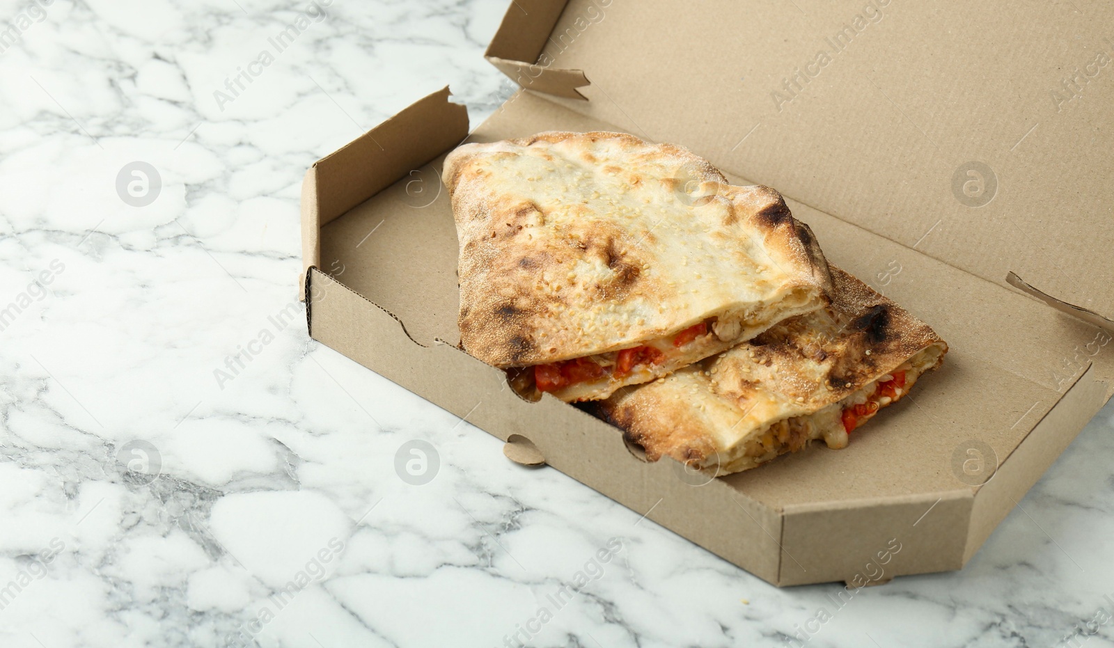 Photo of Halves of tasty calzone with meat, cheese and tomato in cardboard box on white marble table, closeup. Space for text