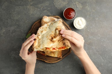 Photo of Woman eating tasty calzone with meat, cheese and tomato at grey textured table, top view