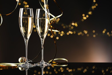 Photo of Champagne in glasses and party streamers on mirror surface against brown background with blurred lights, space for text