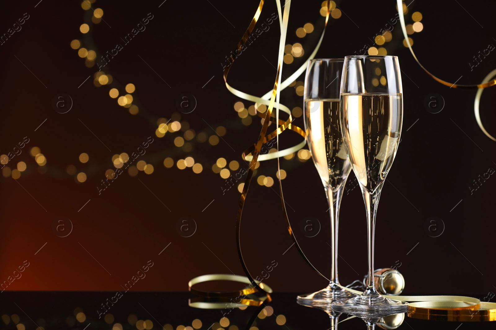 Photo of Champagne in glasses and party streamers on mirror surface against color background with blurred lights, space for text