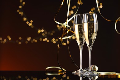 Photo of Champagne in glasses and party streamers on mirror surface against color background with blurred lights, space for text