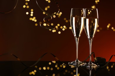 Photo of Champagne in glasses and party streamers on mirror surface against color background with blurred lights, space for text