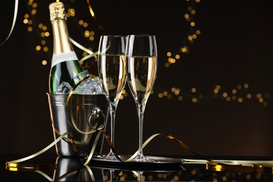 Photo of Champagne and bucket of ice on mirror surface against brown background with blurred lights, space for text