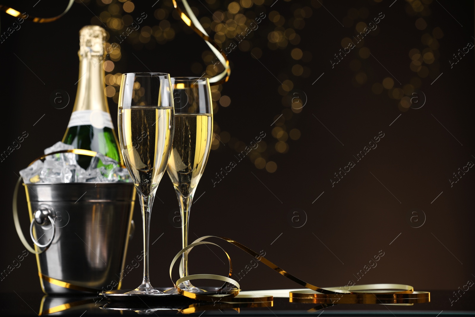 Photo of Champagne and bucket of ice on mirror surface against brown background with blurred lights, space for text