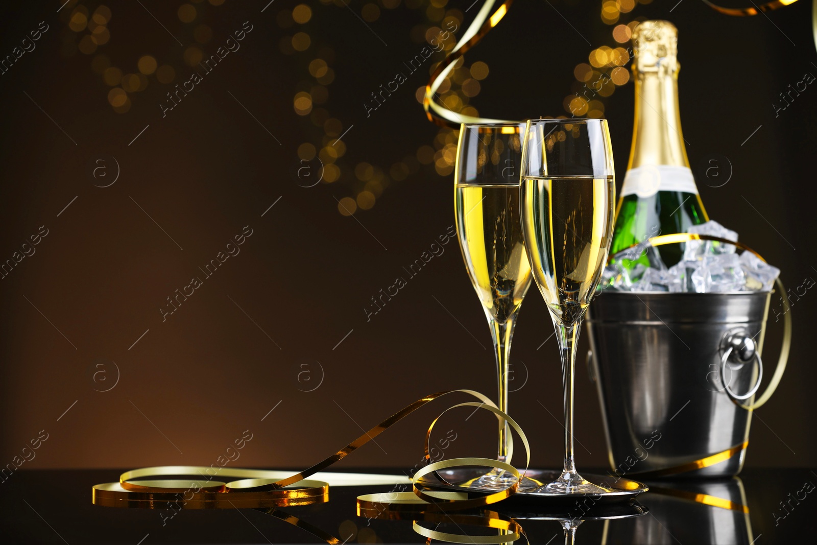 Photo of Champagne and bucket of ice on mirror surface against brown background with blurred lights, space for text