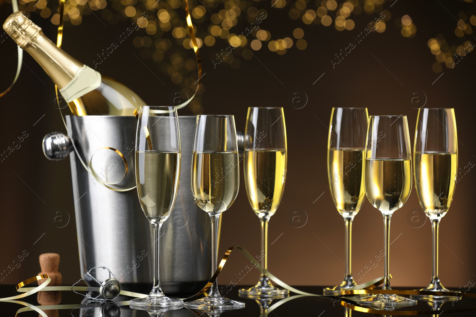 Photo of Champagne in glasses and ice bucket with bottle on mirror surface against brown background with blurred lights