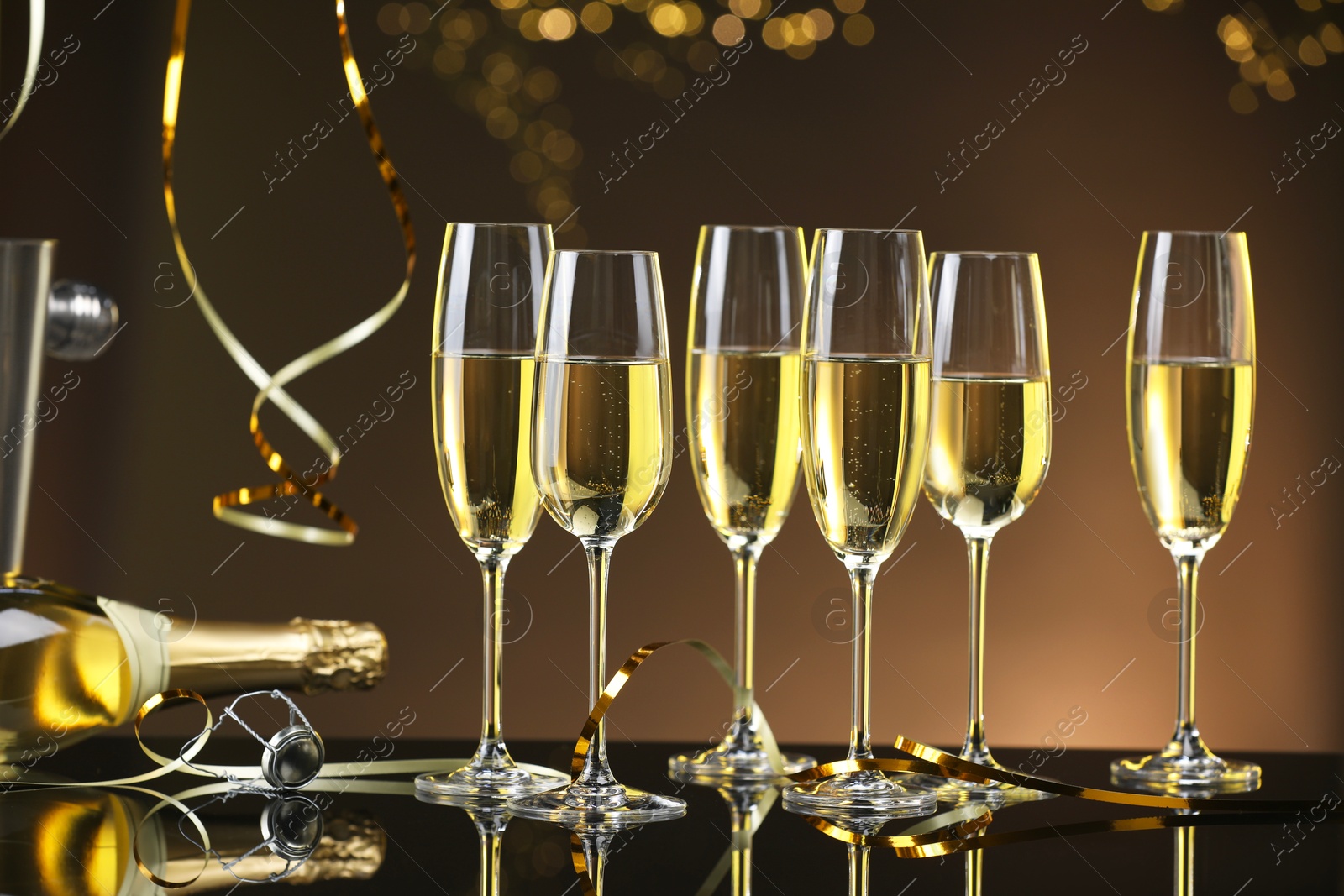 Photo of Champagne in glasses and bottle on mirror surface against brown background with blurred lights