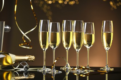 Photo of Champagne in glasses and bottle on mirror surface against brown background with blurred lights