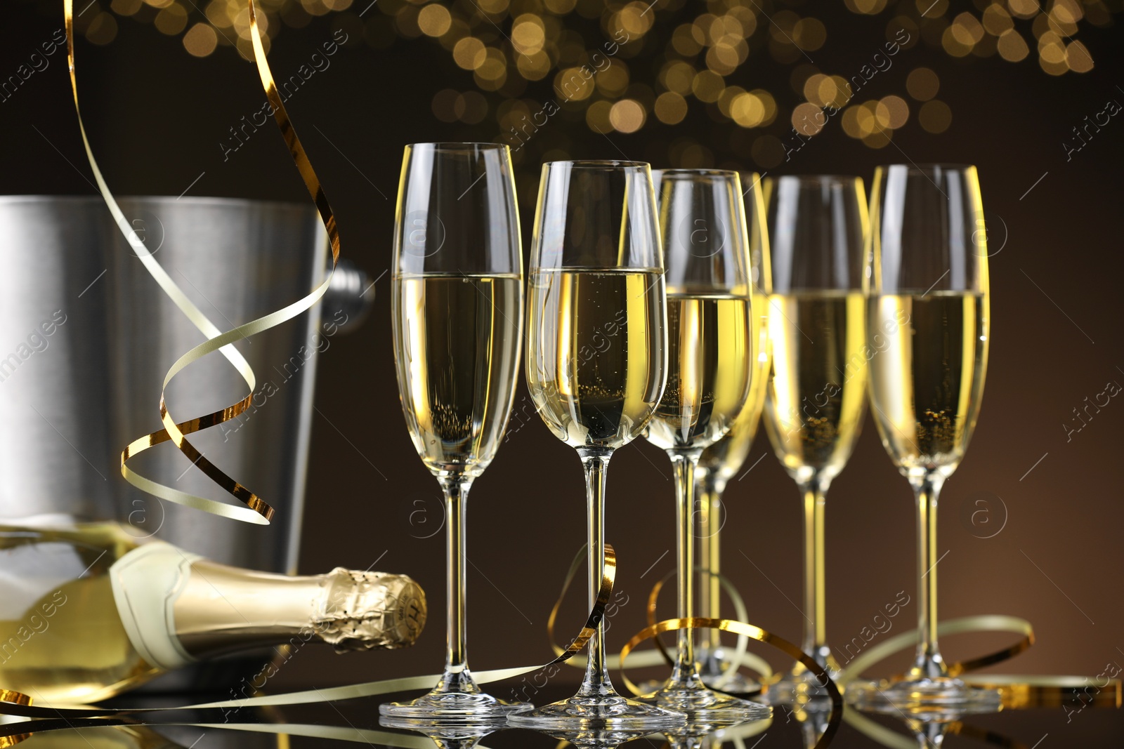 Photo of Champagne in glasses and bottle on mirror surface against brown background with blurred lights