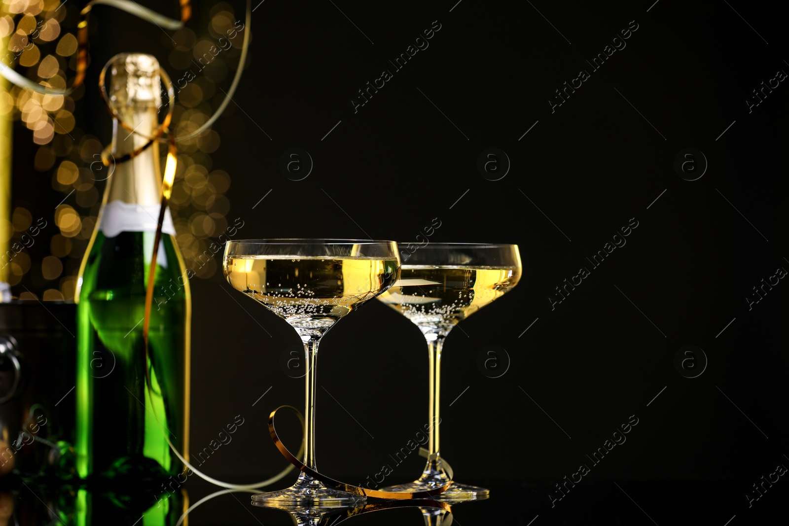 Photo of Champagne in glasses and bottle on mirror surface against black background with blurred lights, space for text